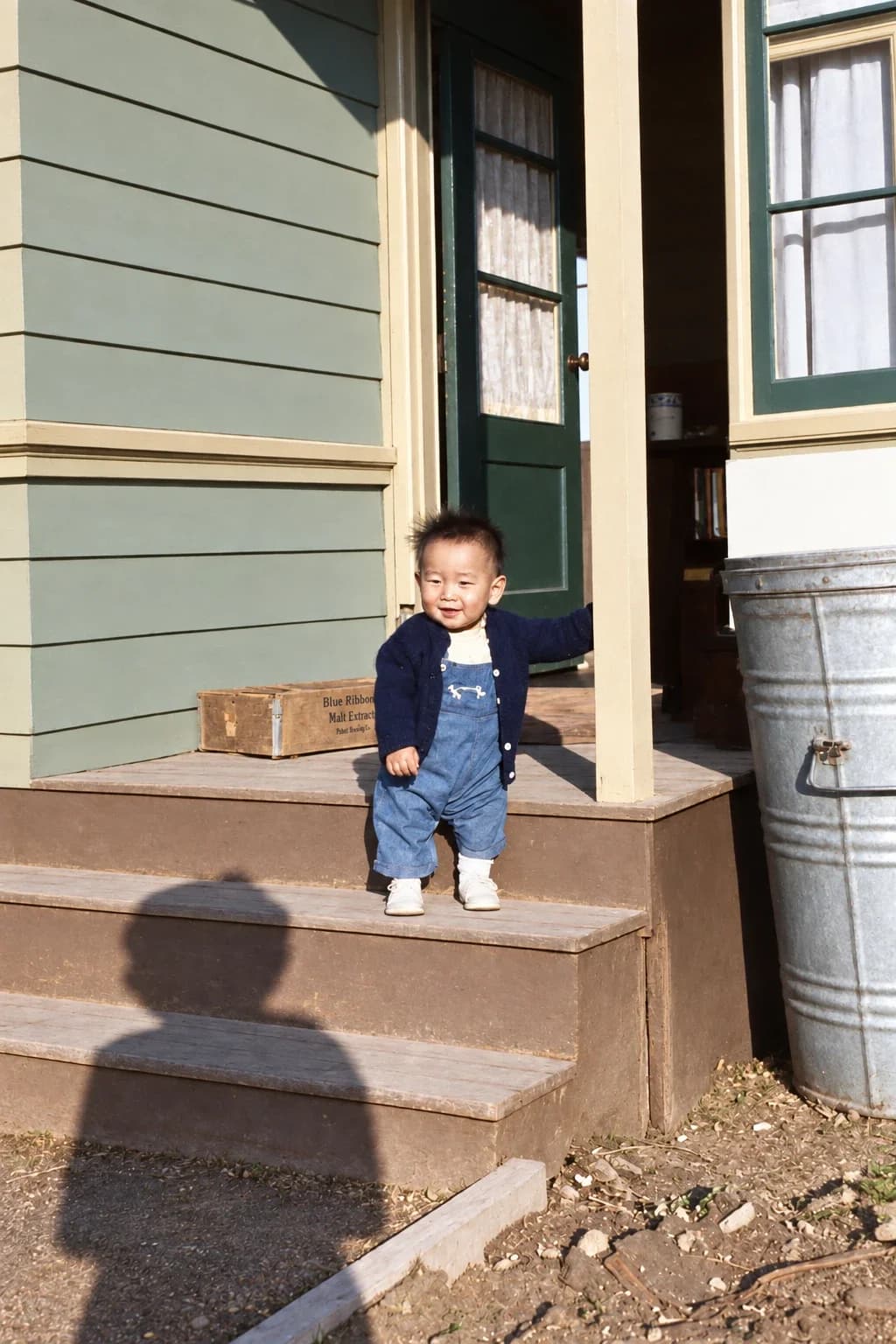 Restored: toddler in a navy cardigan and denim overalls on porch steps, with the photographer's shadow in front